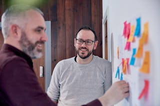 Two men at a whiteboard. One man, in profile, is placing a sticky note on the whiteboard while the other watches, face on.