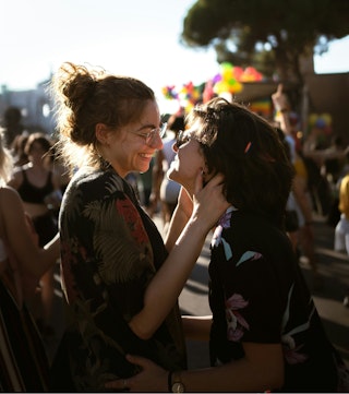Two young women look at each other. They appear to be at an outdoor festival, with the sun setting in the distance. They are smiling