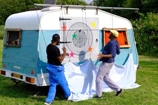 A caravan, with a target on the door. Two men standing in front of it.