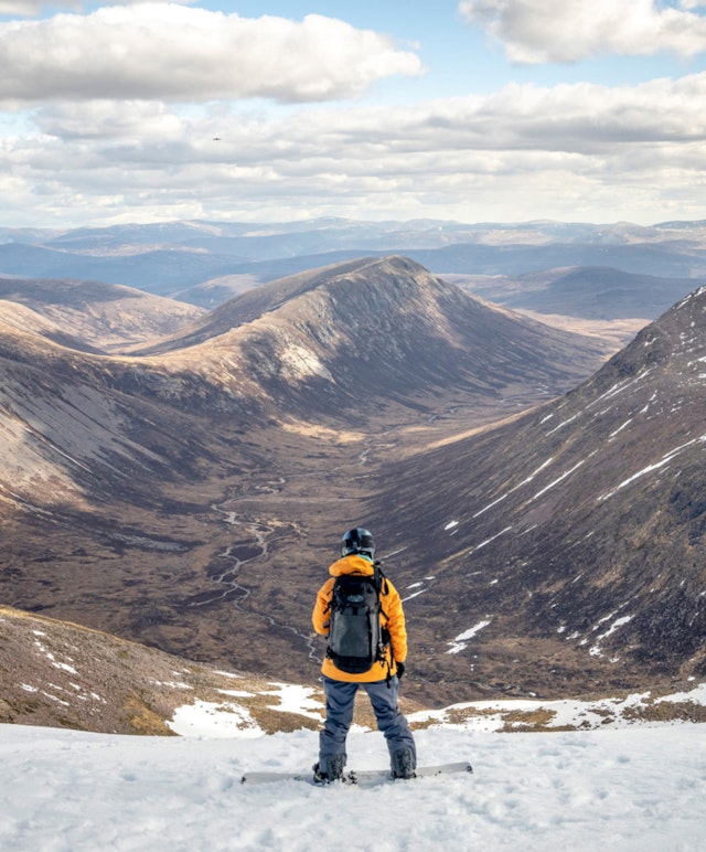 A still from a Patagonia produced video, Thrawn. A snowboarding stands on snow, looking down at snow-less peaks.