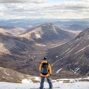 A still from a Patagonia produced video, Thrawn. A snowboarding stands on snow, looking down at snow-less peaks.