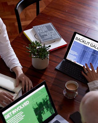 Three people sitting around a wooden table, looking at a tablet and laptop with Backlit Gallery website designs