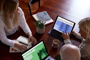 High-level view of three people around a wooden table, looking at digital designs of Backlit Gallery website on a laptop and a tablet