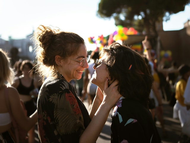 Two young women look at each other. They appear to be at an outdoor festival, with the sun setting in the distance. They are smiling