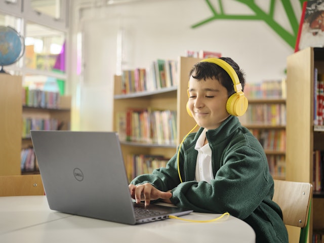 A child with yellow headphones in a library. He is sitting at an open laptop and smiling