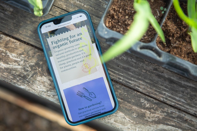 The Garden Organic website on a mobile screen. The mobile is on a garden table, with seedlings behind