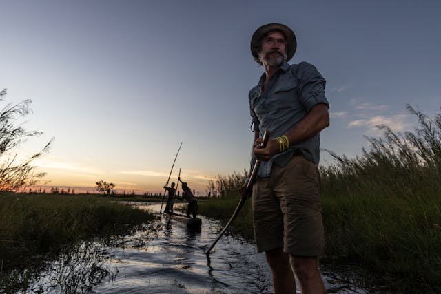 People navigate a shallow waterway with long poles at sunset, surrounded by tall grasses under a clear sky.