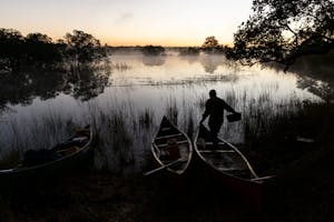A person stands beside two canoes on the shore of a misty river at sunrise, surrounded by reeds and trees.