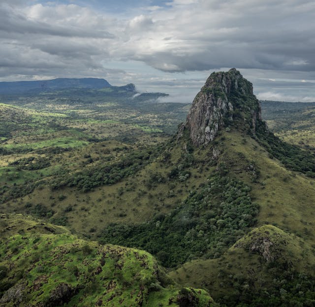 A rocky, steep mountain rises from a green, grassy landscape under a cloudy sky, with distant hills and valleys visible in the background.