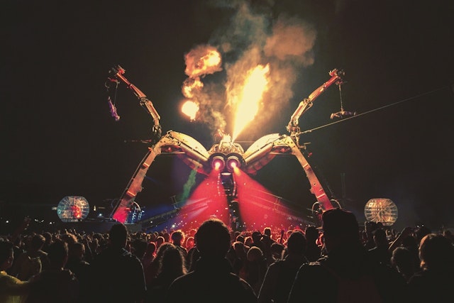 A photograph from Glastonbury Festival - a large robot with two spider-like legs stands over a crowd in the dark. Fire is shooting from it's 'head' and it's 'eyes' light up the crowd with red beams