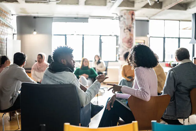Two people sat on chairs chatting in a room that has more people and more chats happening