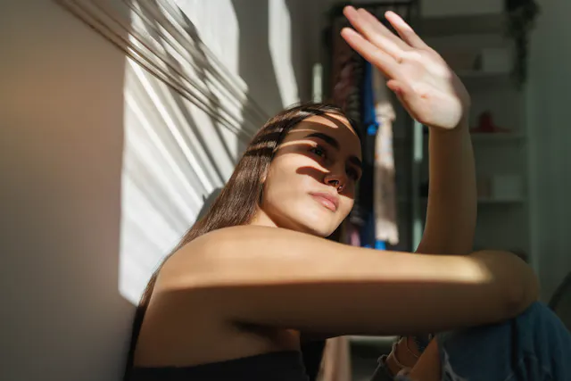 A young woman is sat against a wall, shielding her face from the sun
