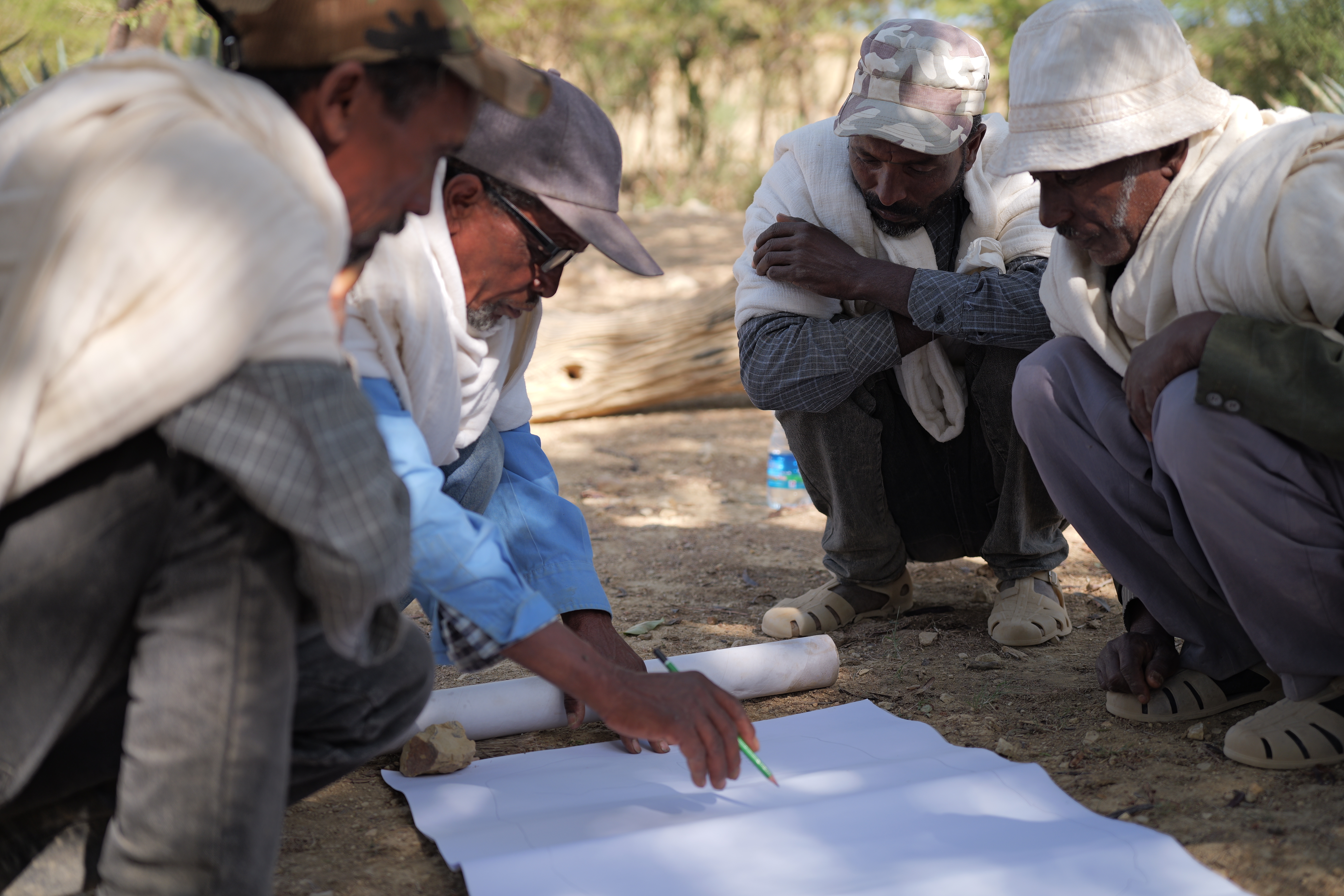 A group of four people crouches over a sheet of paper on the ground and one of them writes on the paper