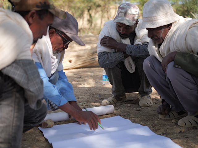 A group of four people crouches over a sheet of paper on the ground and one of them writes on the paper